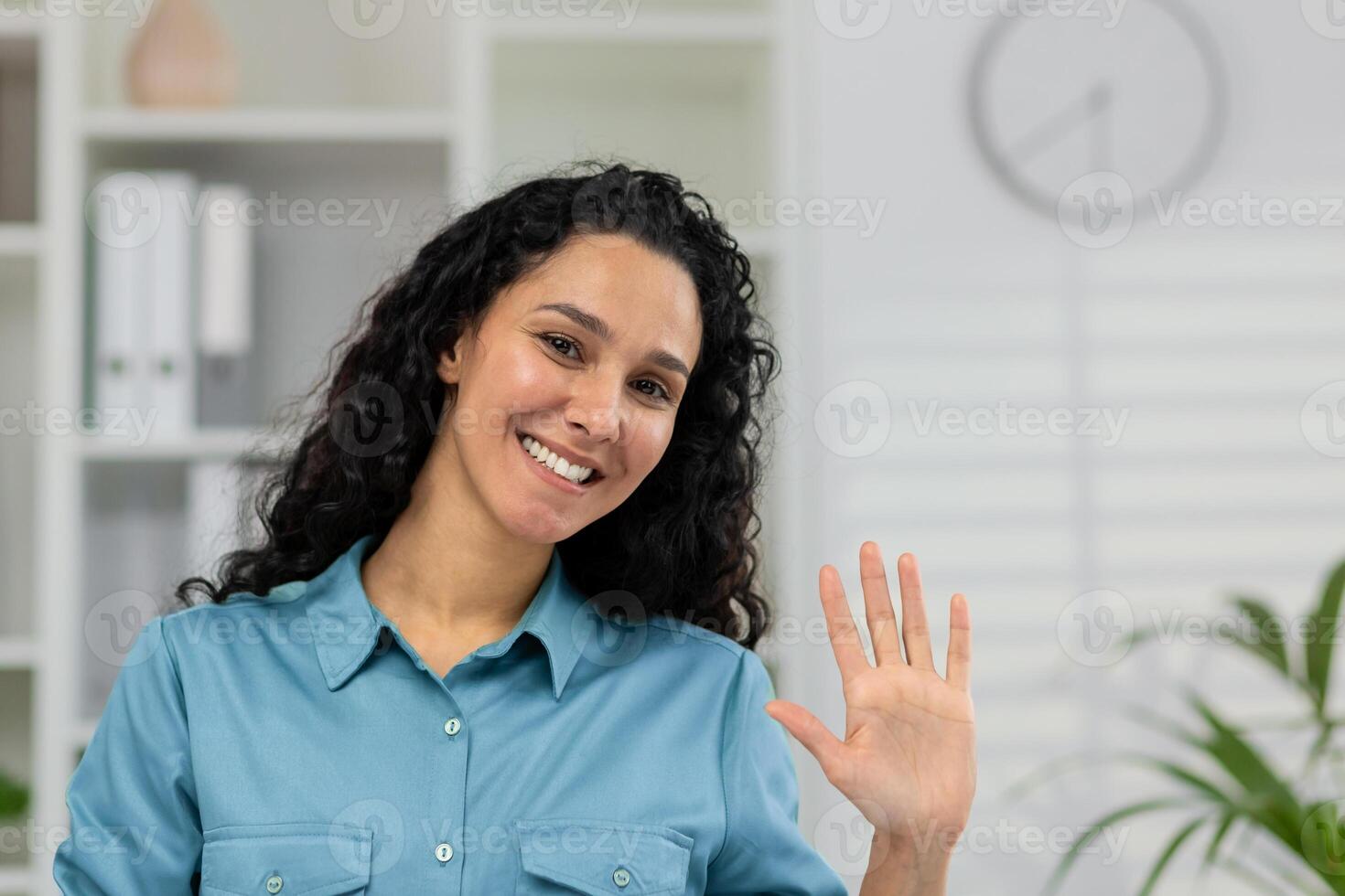 Smiling woman in a casual shirt waving hello during a call, providing an engaging webcam view POV in a home setting. photo