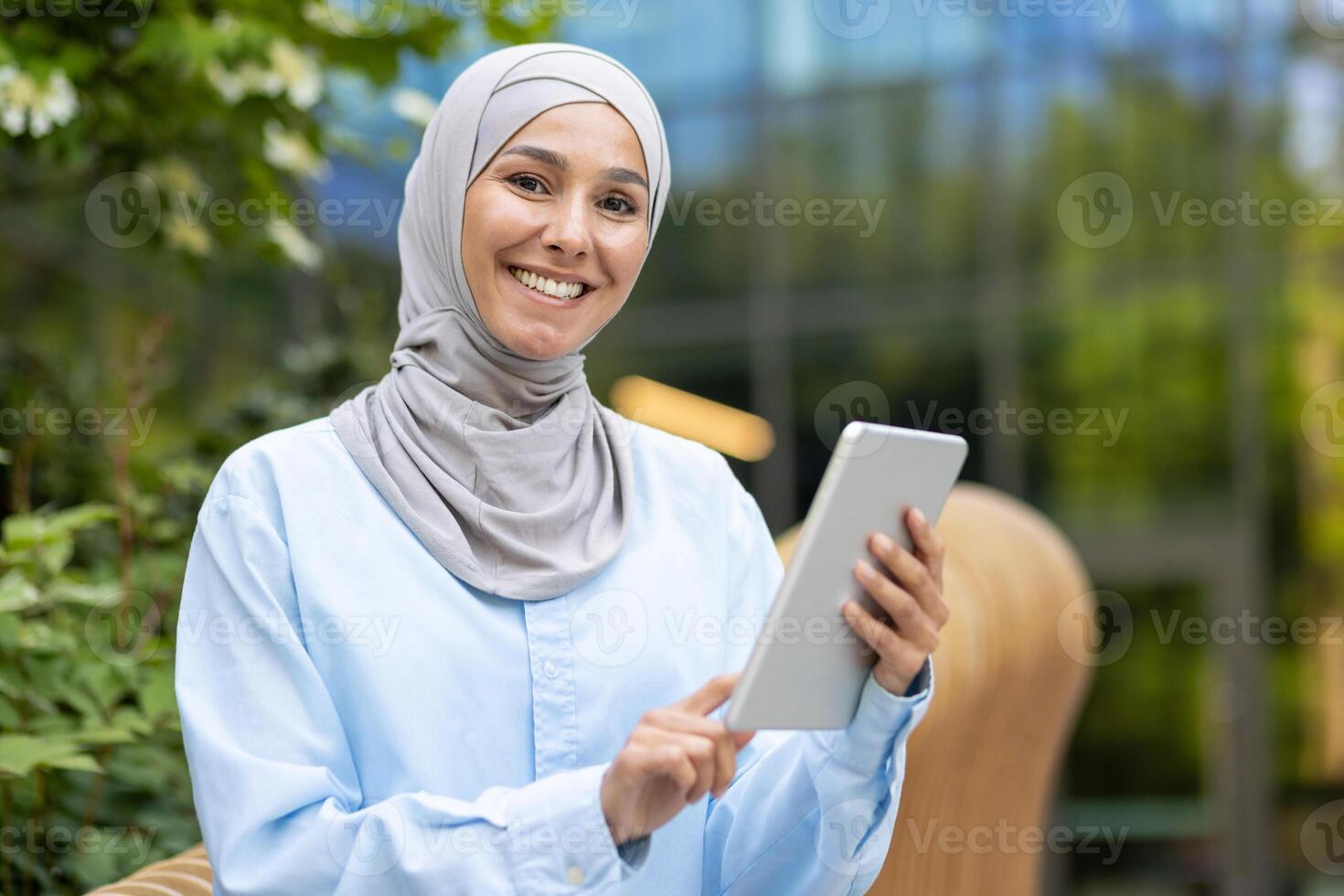A portrait of a happy Muslim woman in a hijab holding a tablet, expressing confidence while standing outdoors with foliage and modern architecture in the background. photo
