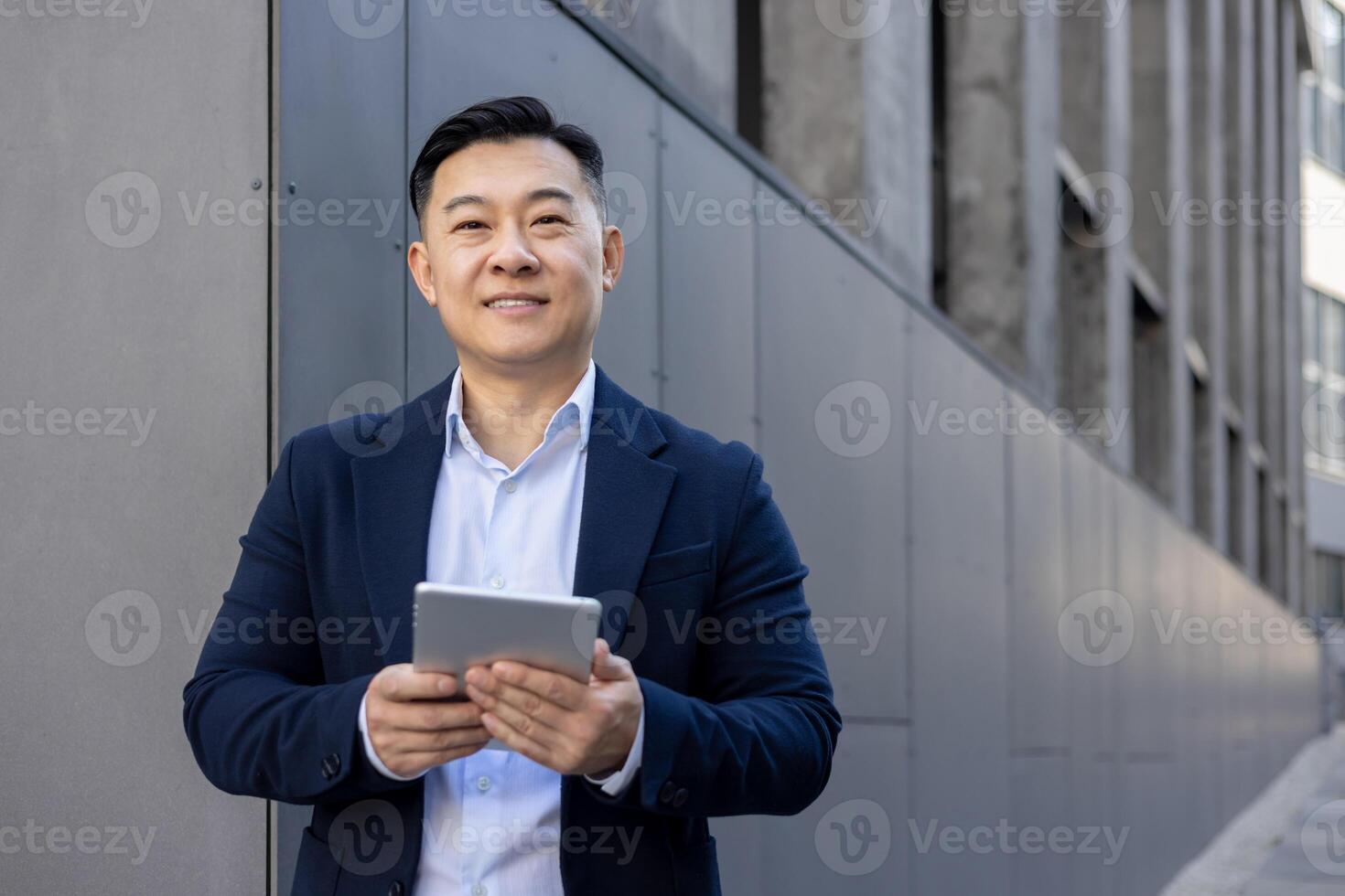 Professional Asian male executive browsing on a digital tablet while standing outdoors with a pleasant expression. photo