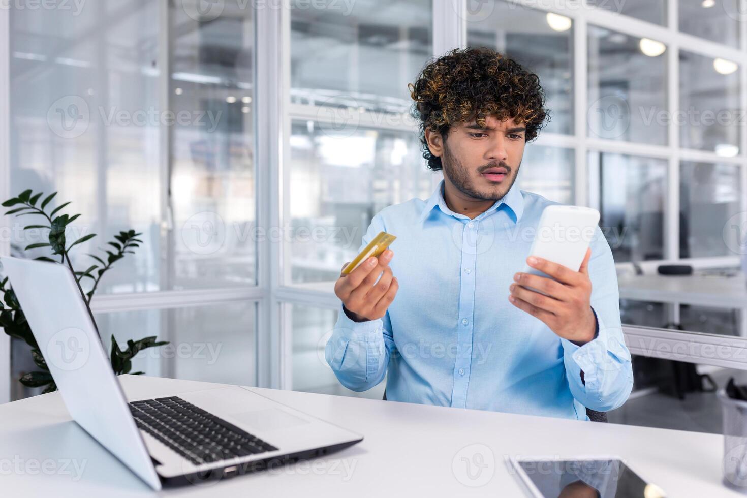 Worried young hispanic man sitting at the desk in the office and looking at the phone upset, holding a credit card in his hands. Unsuccessful online shopping, transfer, blocked account. photo