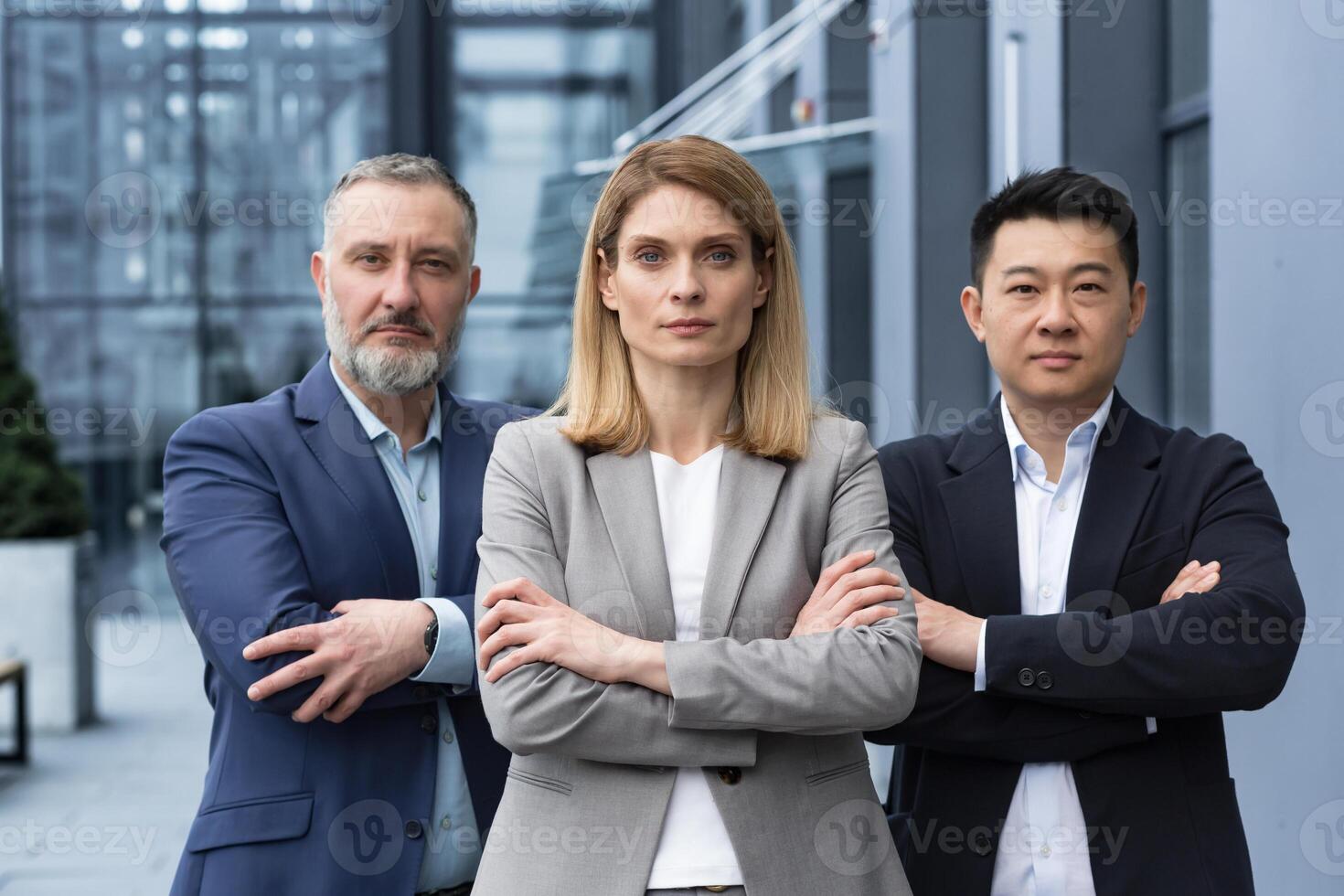 Successful and serious diverse team of three business people, man and woman focused looking at camera with arms crossed, portrait of co-workers outside office building photo