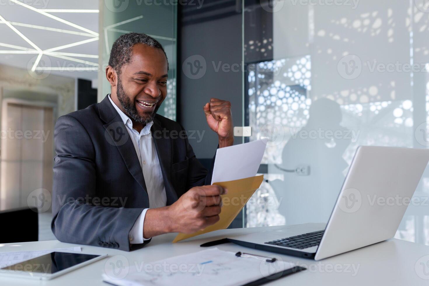 Senior african american man in suit sitting in office at desk and happy. Shows a victory gesture with his hand. He reads the letter in the envelope. Got a job, position, promotion. photo