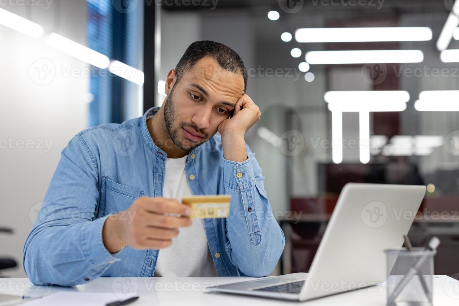 A man appears worried as he holds a credit card and stares at his laptop screen, possibly dealing with financial issues. photo