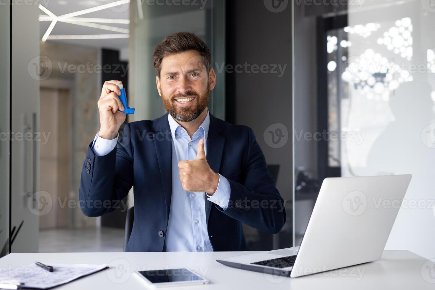 retrato de un joven empresario sentado a un escritorio en el oficina en un traje, participación y demostración un asma y alergia rociar en su mano, sonriente y mirando a el cámara. foto