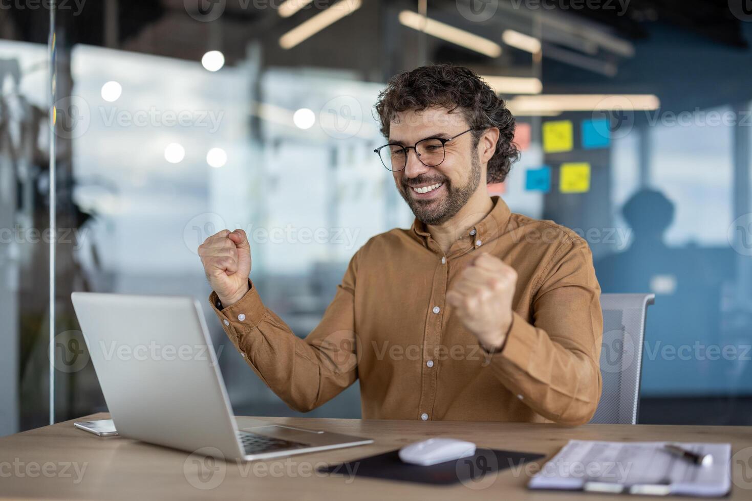 Joyful male professional in a casual shirt jubilantly celebrating a work victory or success at his office workspace with a laptop. photo