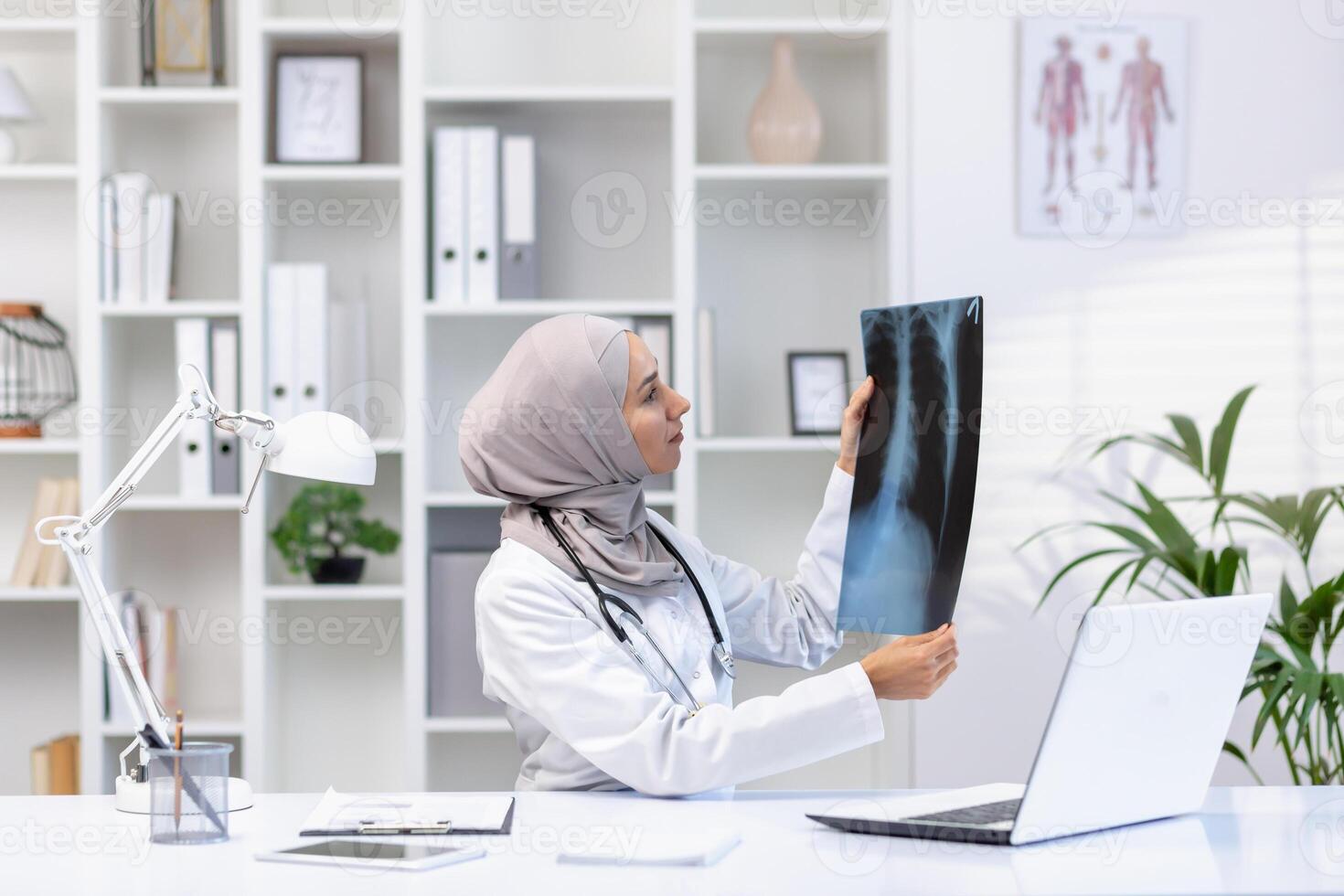 enfocado musulmán mujer médico con estetoscopio comprobación radiografía de livianos mientras sentado a hospital gabinete con ordenador portátil. calificado neumólogo en blanco médico uniforme haciendo diagnóstico para paciente. foto