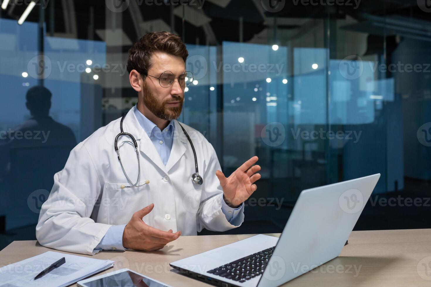 Online consultation of mature serious doctor with beard, man in medical gown using laptop for call, explaining treatment instructions to patient, working inside modern clinic in the evening. photo