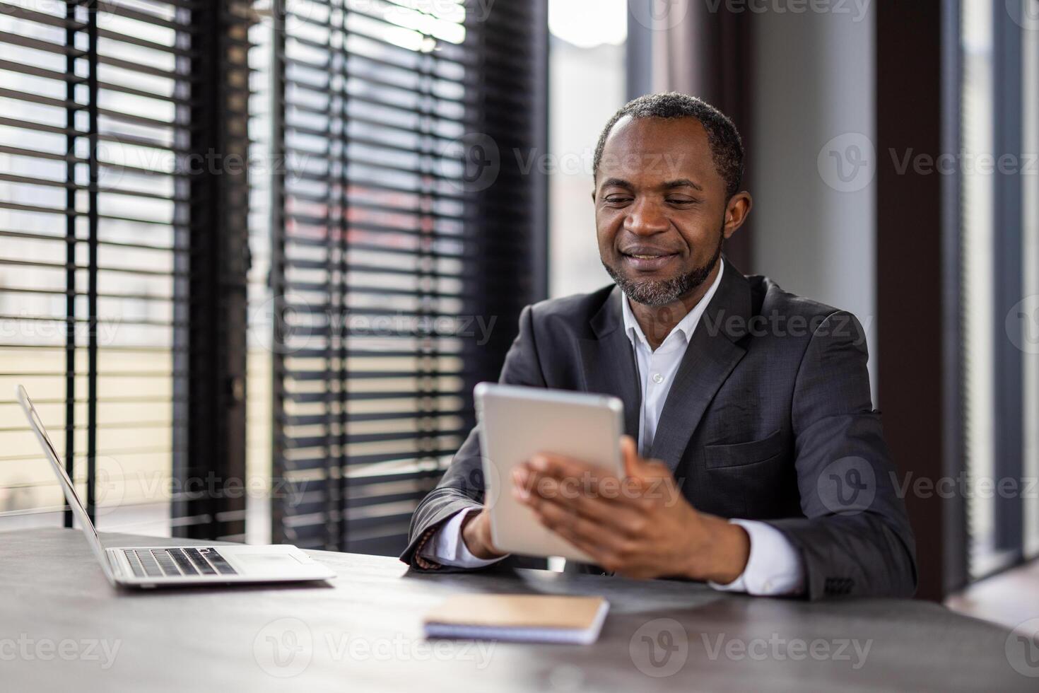 sonriente africano americano empresario trabajando en moderno oficina con tableta y computadora portátil, representando profesionalismo y éxito. foto