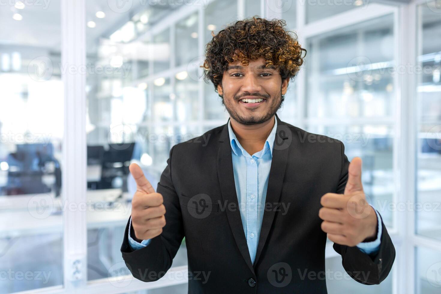 retrato de exitoso Hispano empresario jefe, gerente en negocio traje y barba mirando a cámara y sonriente en pie cerca ventana, demostración pulgares arriba, firmar de éxito y lograr objetivos. foto