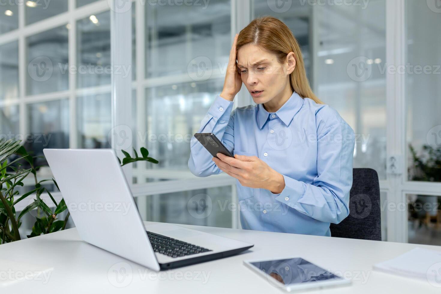 Worried freelancer grabbing head in frustration while checking time on cellular phone and sitting in coworking. Stressed female administrator not fitting in time frame for project completing. photo