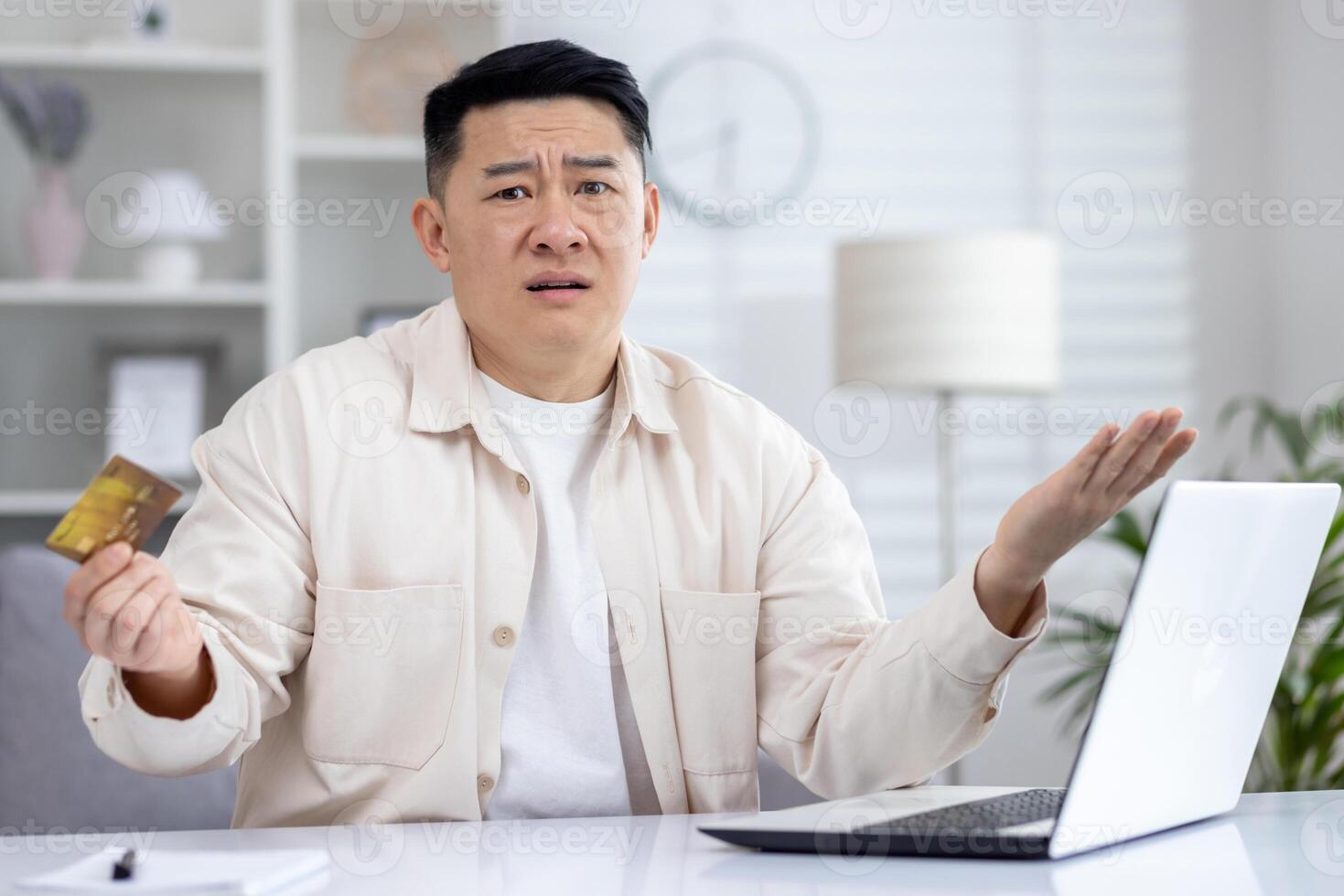Confused asian man sitting in light office at table in front of laptop, holding card in hand, upset about failed online shopping, broken computer, failed online transaction. photo