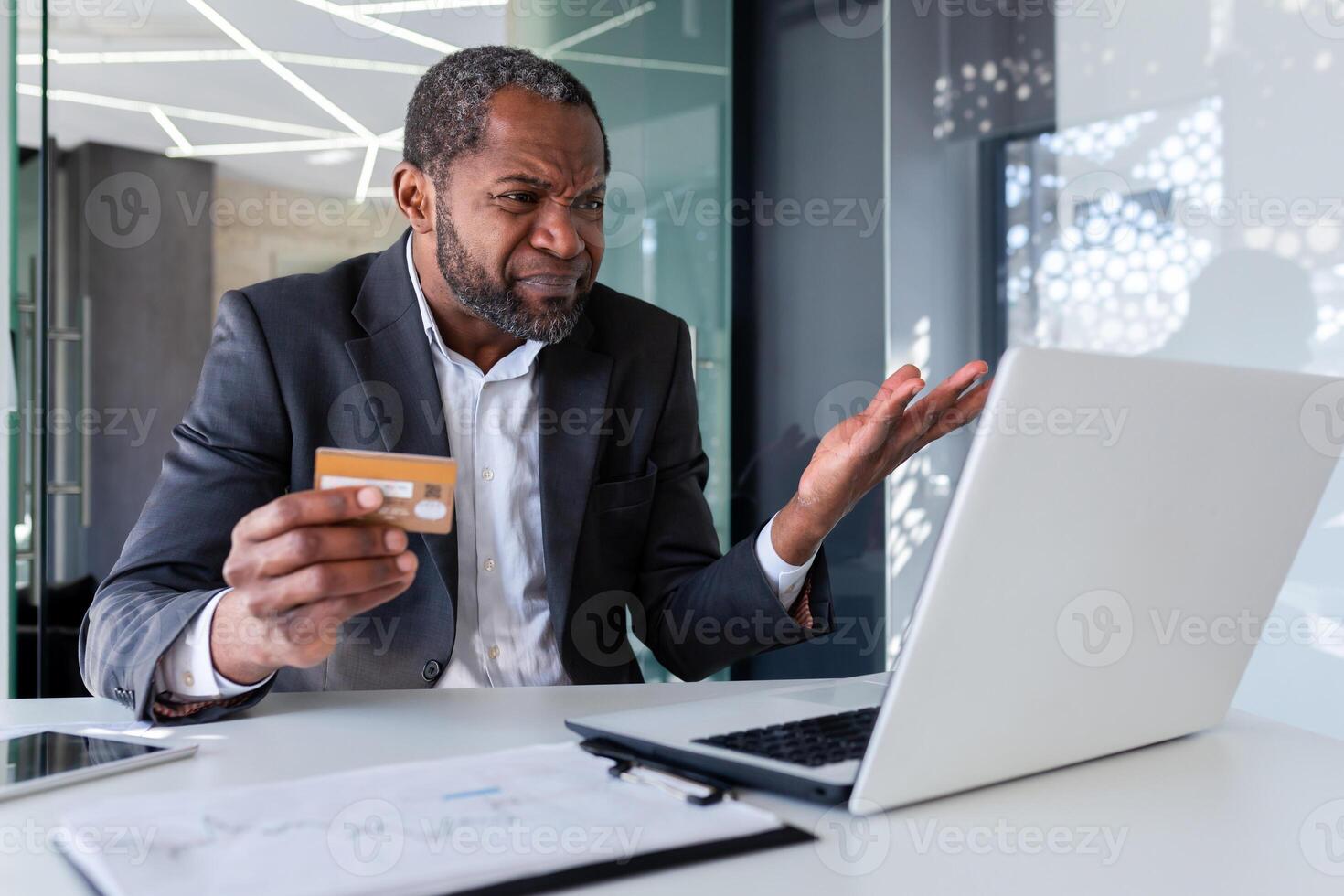 Upset and worried businessman at workplace, man cheated received online money transfer error, african american man unhappy sitting with laptop inside office holding bank credit card. photo