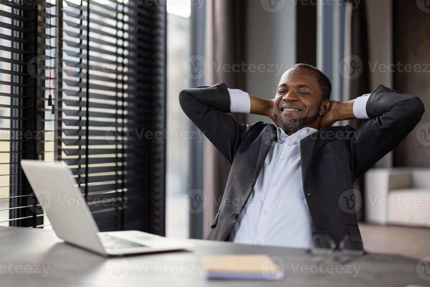 Satisfied African American entrepreneur in a stylish suit takes a peaceful break at his workspace with a laptop. photo