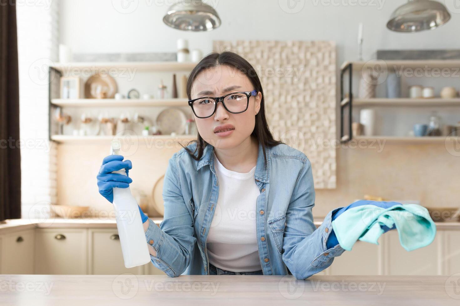 Tired young beautiful Asian woman cleaning at home. Wipes the table with a rag in rubber gloves and a bottle of spray. He looks at the camera, spreads his hands. photo