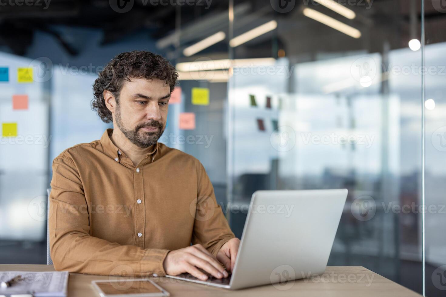 Focused professional man using a laptop in a contemporary office setting with glass partitions and sticky notes in the background. photo