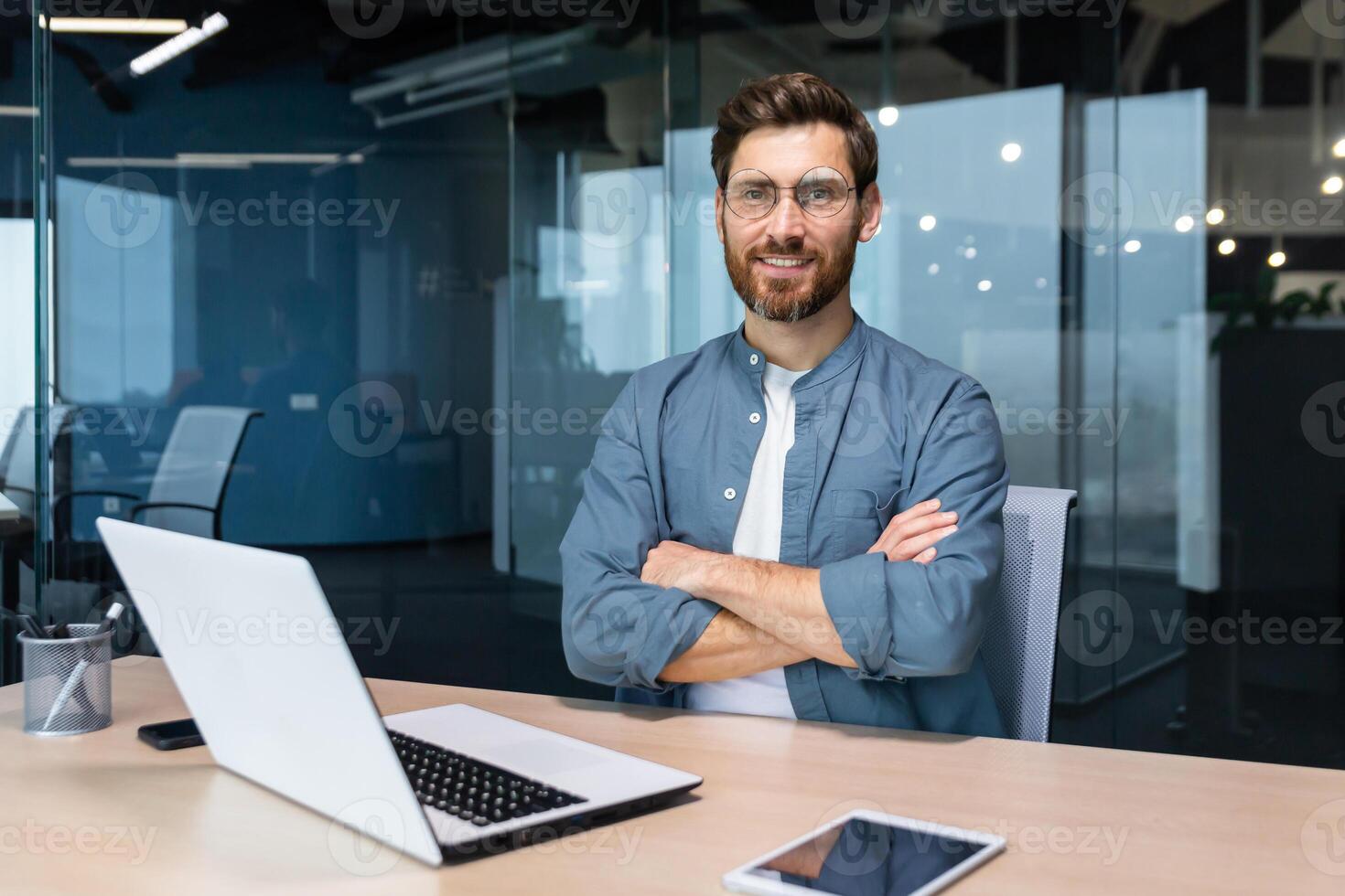 Portrait of mature businessman with beard, man in shirt smiling and looking at camera sitting at desk with crossed arms inside modern office building, using tablet and laptop at work. photo