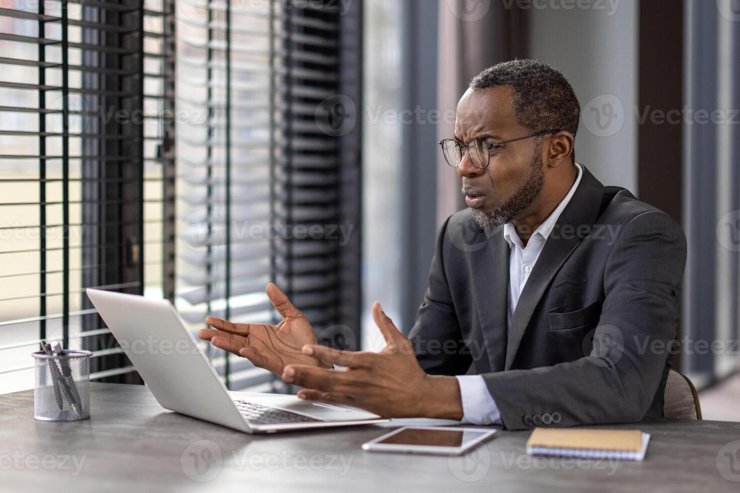 Clueless business person spreading hands to computer while working by table with tablet and notebook. Mature man in jacket trying solving company processes and feeling desperate about new tasks. photo