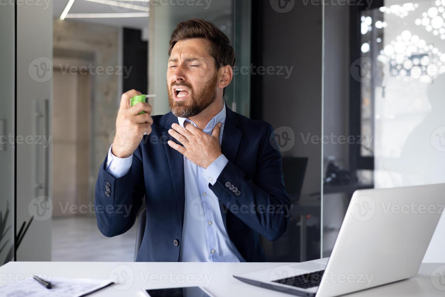 A young male businessman suffers from a severe sore throat, sits at a desk in the office and snorts a medicine aerosol into his mouth. photo