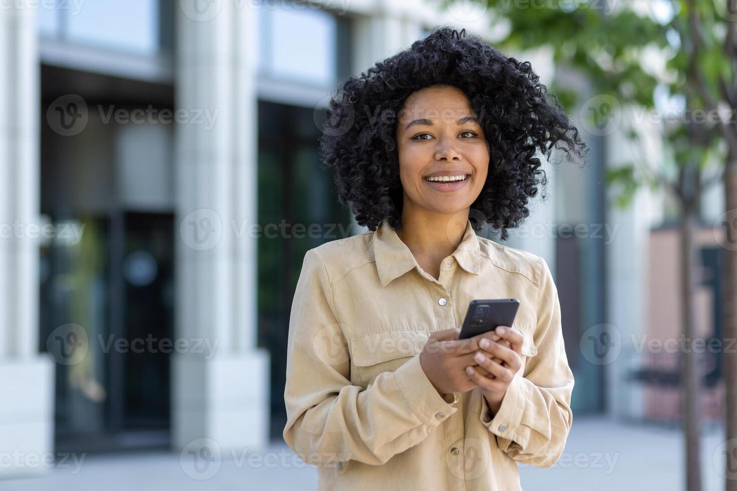 Portrait of young beautiful African American female student outside campus walking with phone in hands, business woman in casual shirt smiling and looking at camera while typing message and call. photo