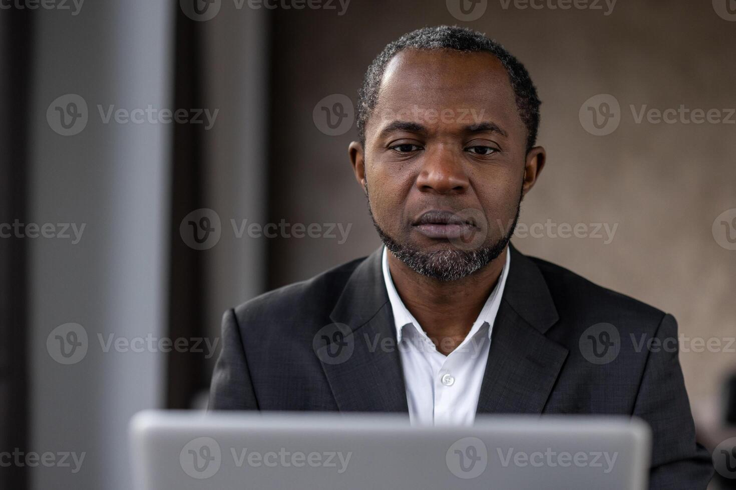 Portrait of concerned diverse man looking attentively into monitor of computer on blurred background. Strict employer in shirt checking productivity of marketing department remotely from cabinet. photo