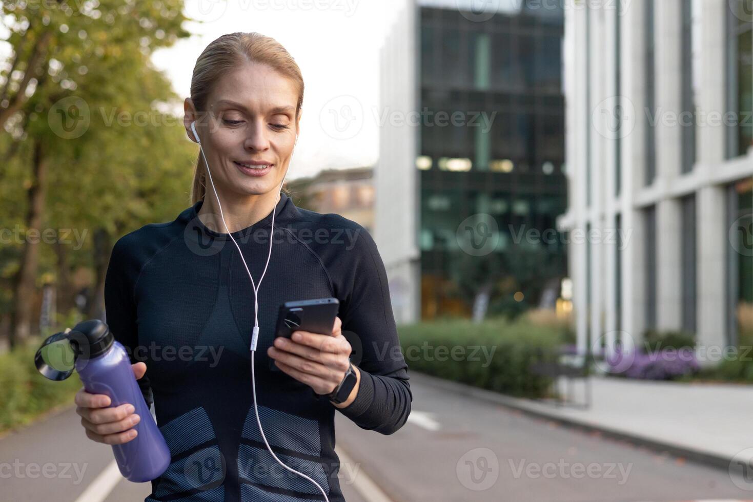 Focused female runner choosing songs on her phone with earphones, holding a water bottle post jog in a city setting. photo