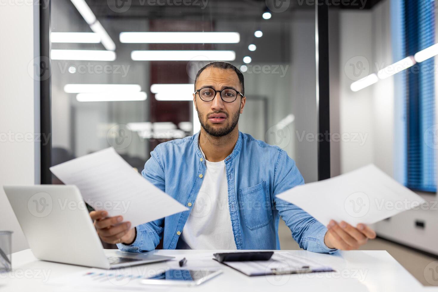 Perplexed Indian man working as an office worker, experiencing confusion while reviewing documents, with a laptop on his desk. photo