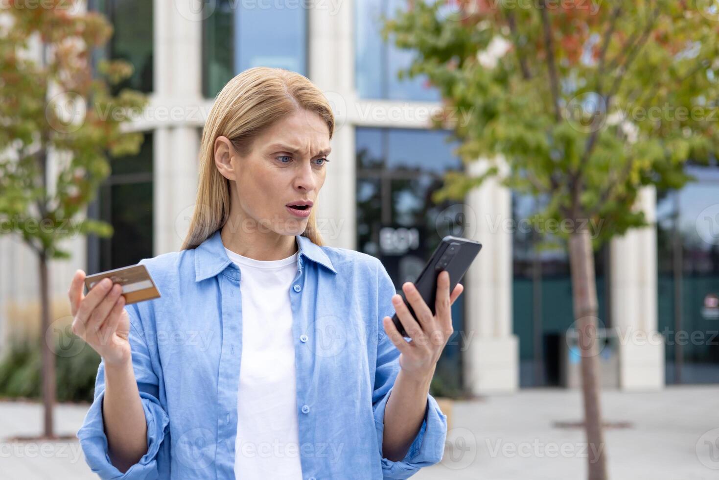 Deceived woman outside office building denied money transfer, mature female worker with phone and bank credit debit card standing, upset and unhappy blonde. photo