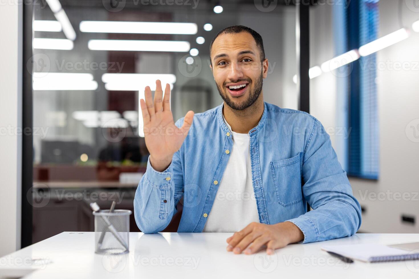 Cheerful Indian businessman in a casual blue shirt engaging in a call from his modern office, greeting with a wave. photo