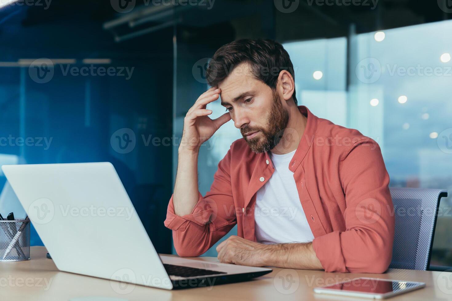 Sad and thinking programmer in red shirt working on laptop inside office, mature man with beard. photo