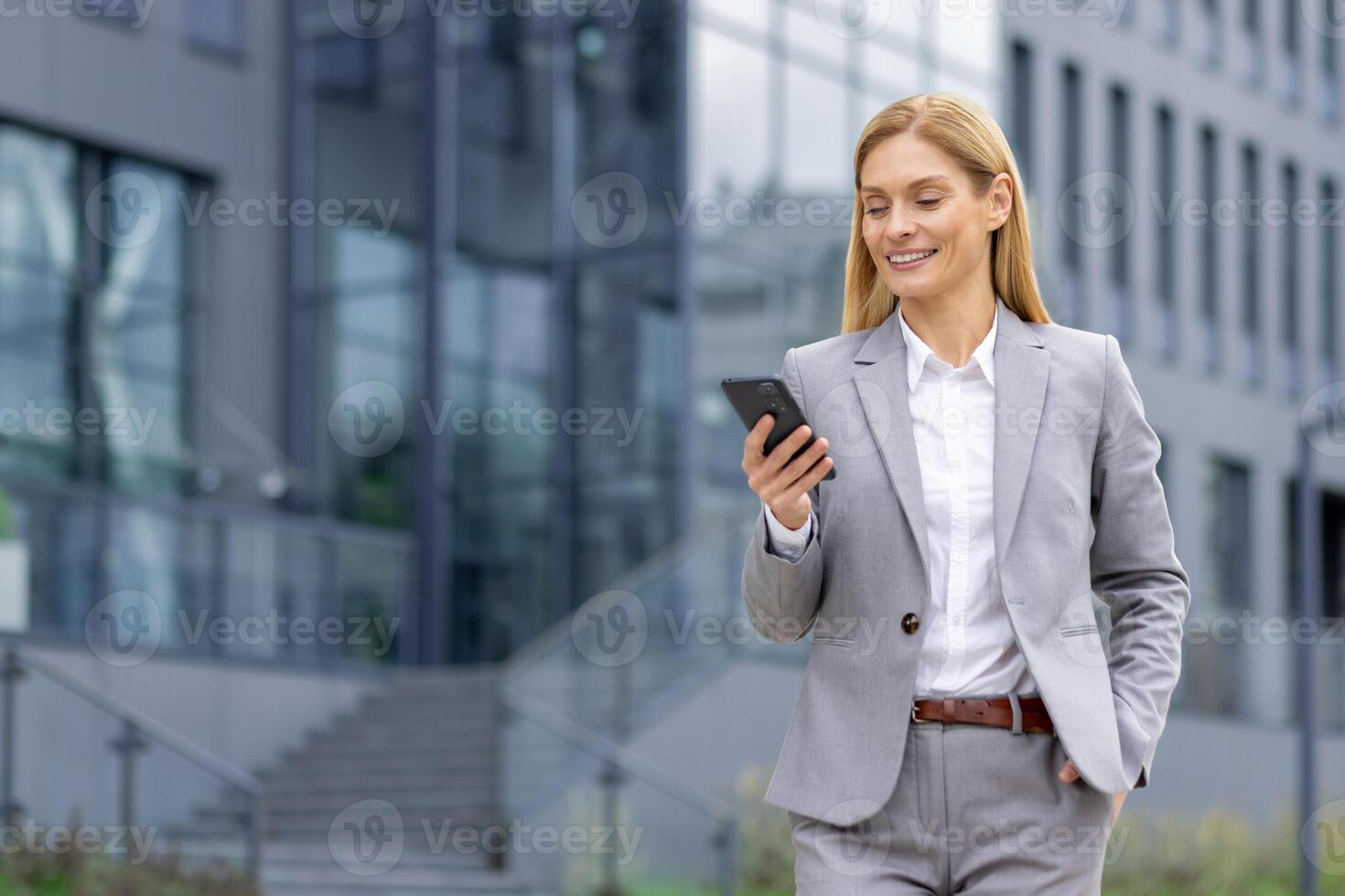 Successful woman boss walks outside office building outdoors, in business suit, businesswoman uses app on phone, smiles contentedly reads internet pages, browses news, writes text message, call. photo