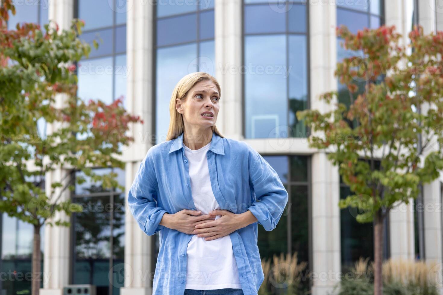 maduro mujer en un camisa es caminando en el ciudad, un trabajador tiene un grave estómago dolor, un rubia mujer de negocios en casual ropa es enfermo, masajear su manos con su manos fuera de un oficina edificio. foto