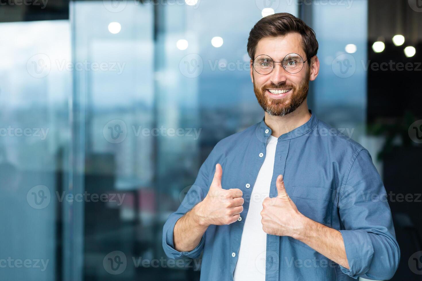 Portrait of businessman in casual shirt, man with beard and glasses smiling and looking at camera showing thumbs up affirmative, business owner working inside office. photo