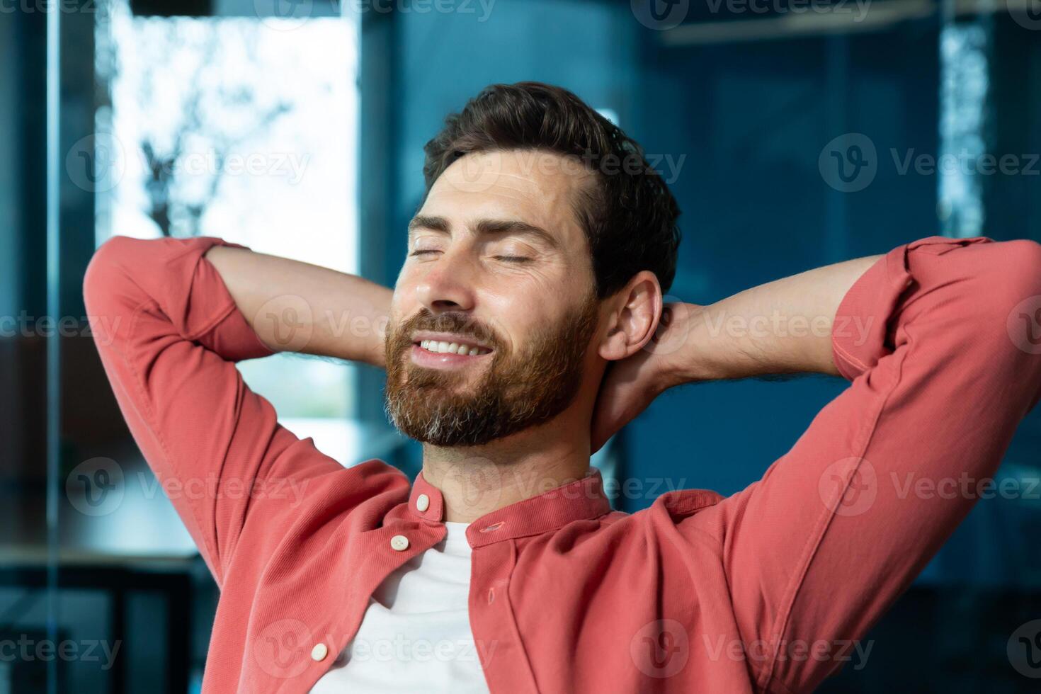Successful businessman in office in a red shirt rests with his hands folded head with closed eyes, mature man with a beard works with a laptop at the workplace inside, dreams of future achievements. photo