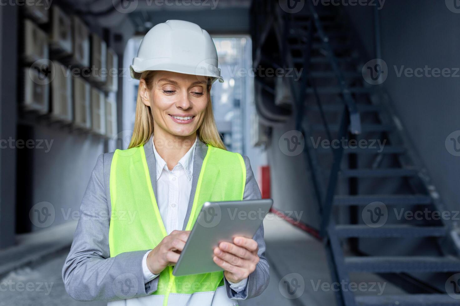 A smiling female engineer in a hard hat and safety vest busy with a tablet on a construction site. photo