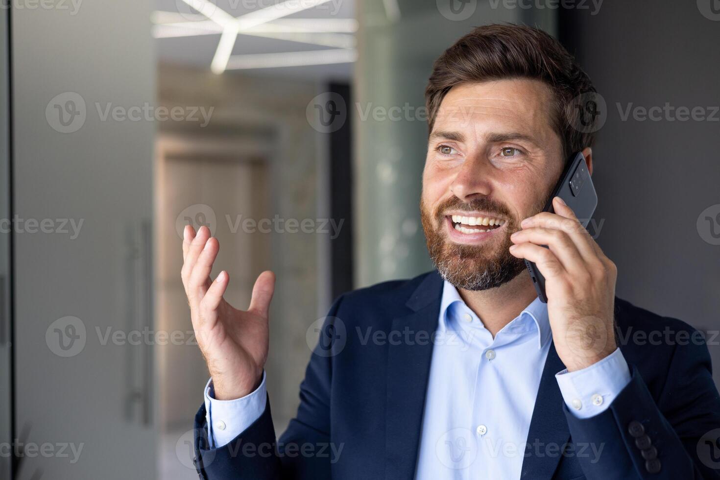 Close-up photo of successful mature businessman talking on the phone, man in business suit smiling contentedly at workplace inside office, consulting clients remotely.