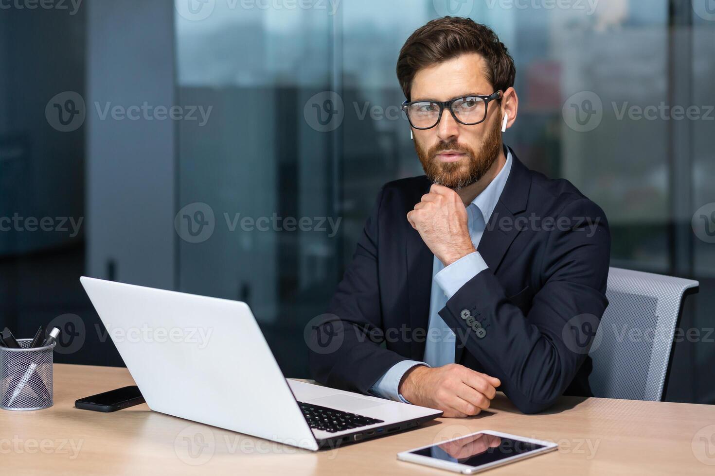 grave pensando maduro empresario trabajando dentro moderno oficina edificio, mayor jefe en negocio traje y lentes trabajando sentado a computadora portátil, hombre con barba pensando acerca de futuro estrategia plan. foto