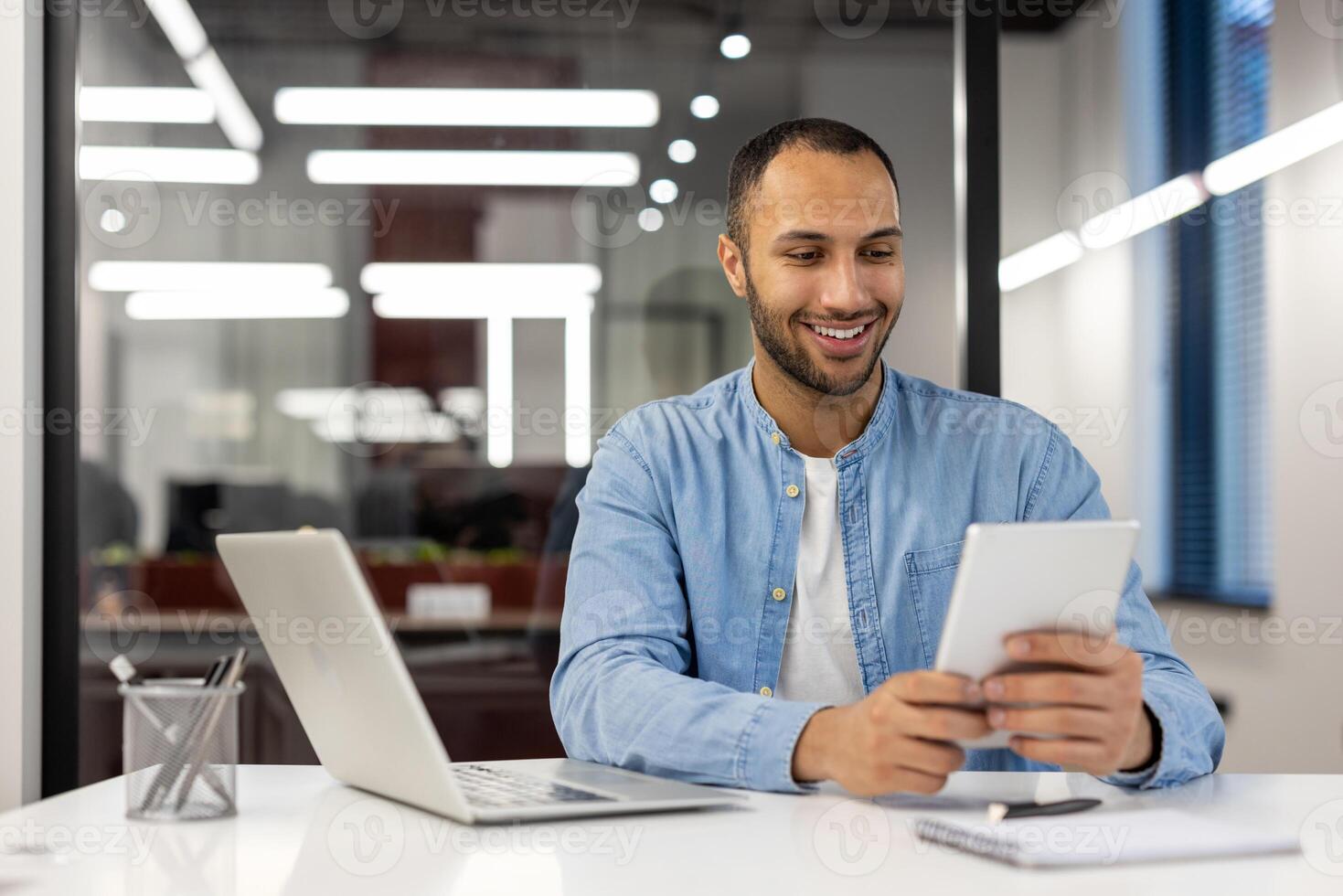 A focused professional multitasking with a tablet and laptop at his sleek workspace in a contemporary office. The image conveys productivity, technology, and modern business environment. photo