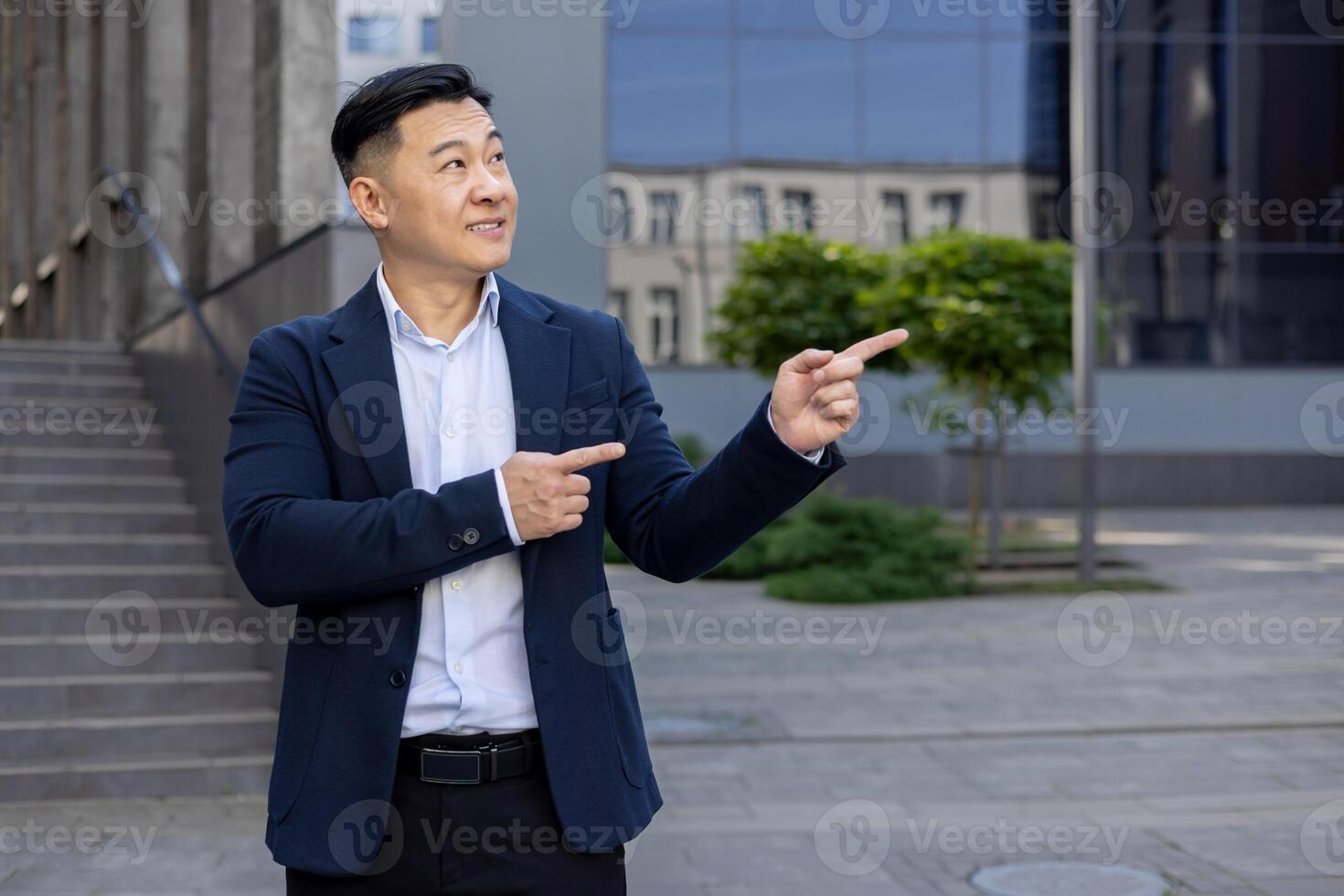 Asian businessman in suit pointing with both hands to side on a sunny day, standing outside with buildings in background. photo