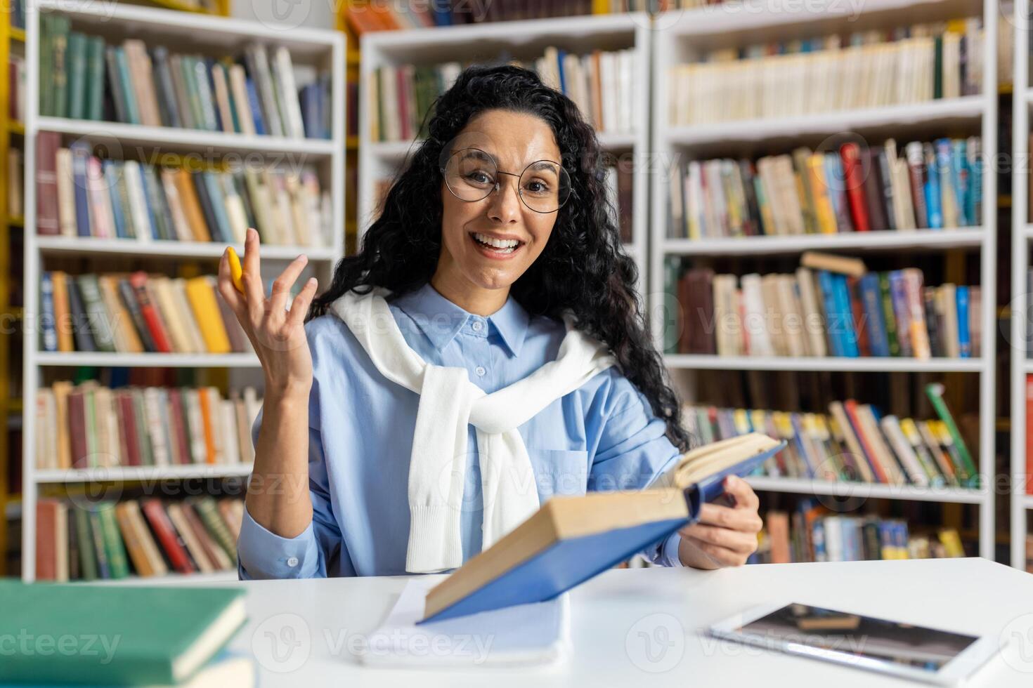 A cheerful female librarian, dressed in a light blue shirt and sweater, explaining from a book amongst shelves filled with diverse books. Portrays positive education and learning environment. photo