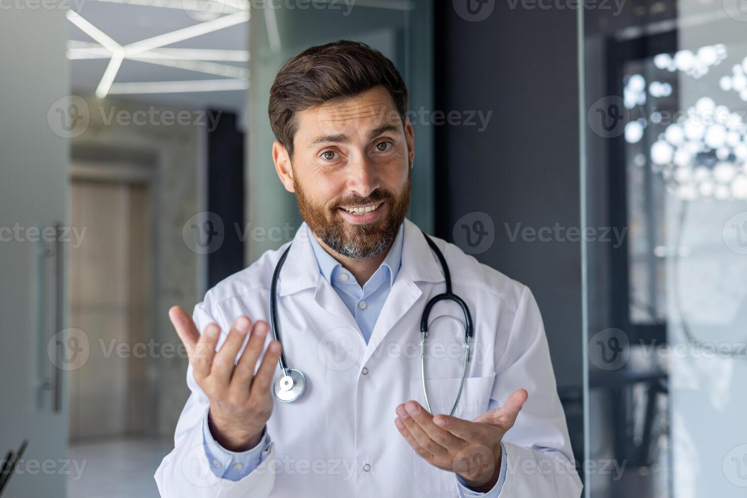 Close-up portrait of a young smiling male doctor who is in the hospital and talking via link, conducting an online consultation and meeting, gesturing with his hands. photo