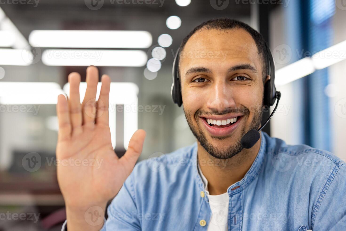 Close-up portrait of a young hispanic man wearing a suit sitting in the office, smiling at the camera and waving. photo