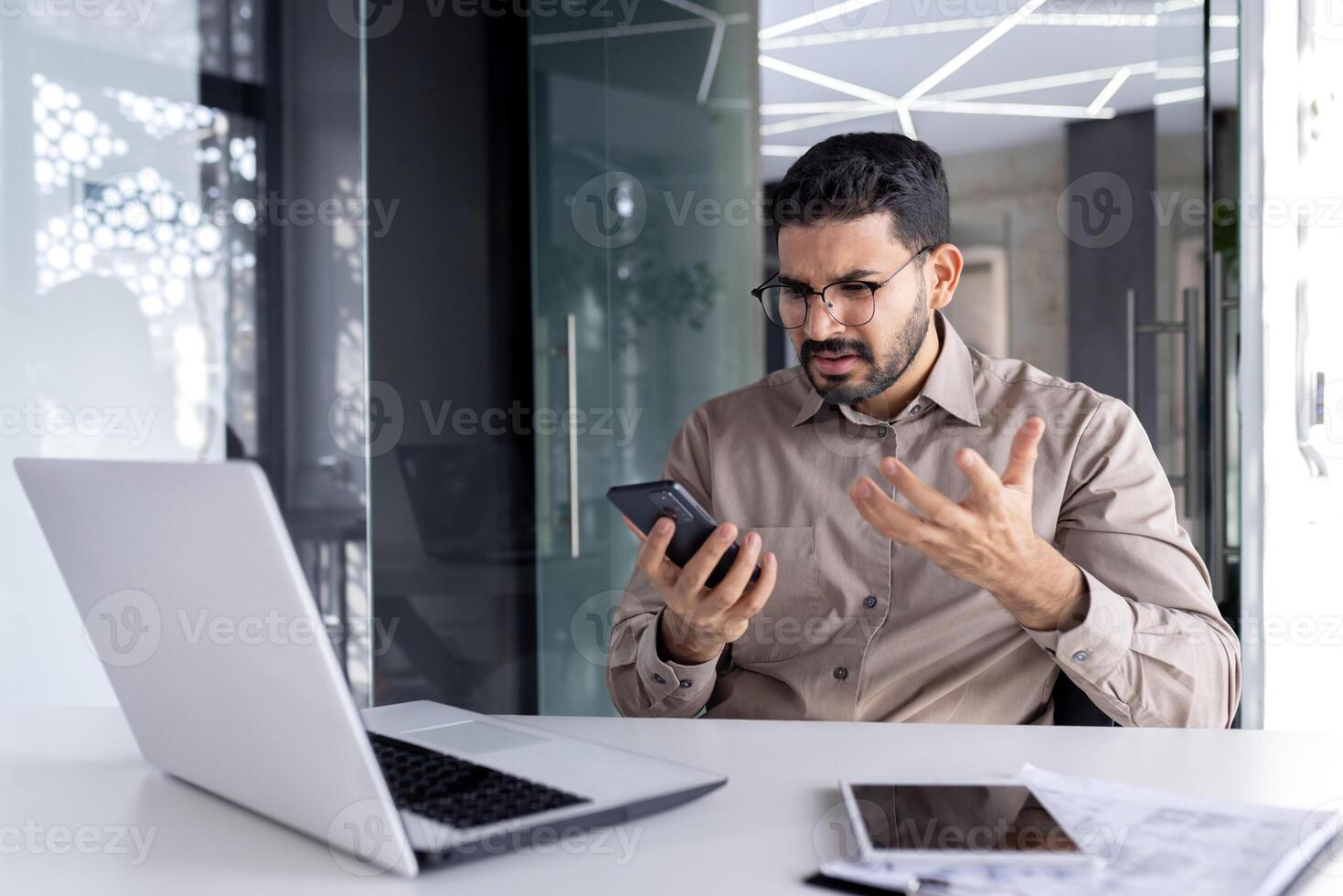Upset sad man at workplace inside office, businessman uses phone reads bad news received by e-mail, employee disappointed holds smartphone in hands browses internet sites and social networks. photo