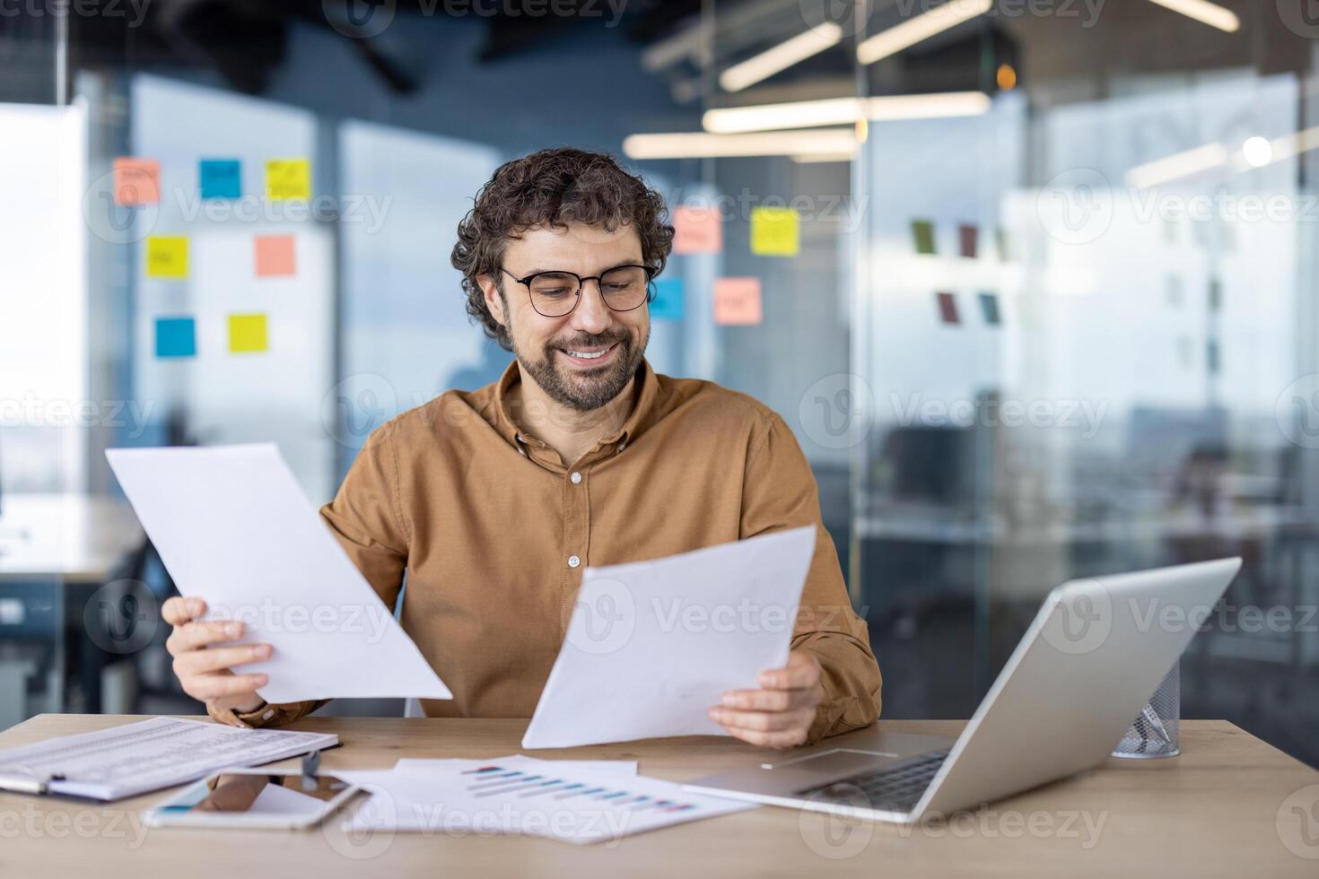 Happy businessman checking documents with records while sitting by table with portable computer and tablet. Satisfied person calculating financial indicators and comparing expenses and income. photo