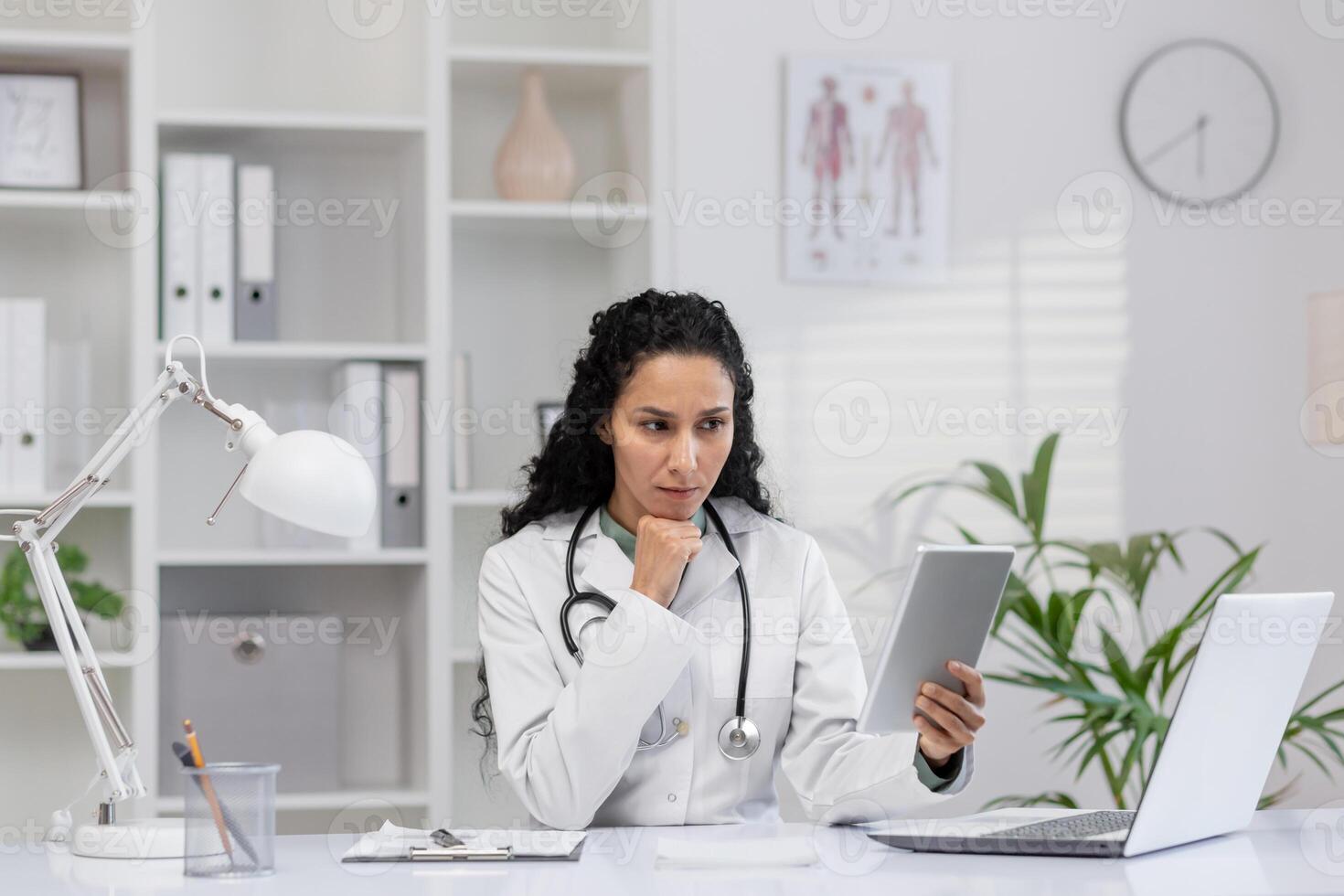 Focused female doctor in a white coat sitting at her office desk, examining medical information on a tablet with a laptop nearby in a well-lit clinic. photo