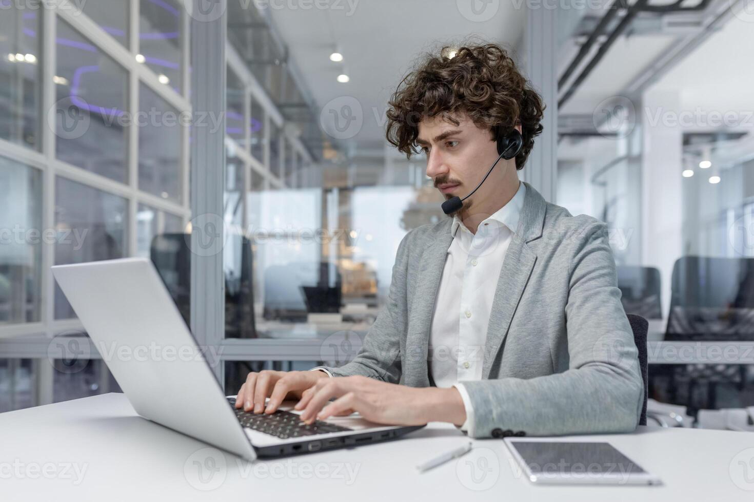 A serious young businessman man in a headset is sitting in the office at a desk and is concentrating on working on a laptop. photo