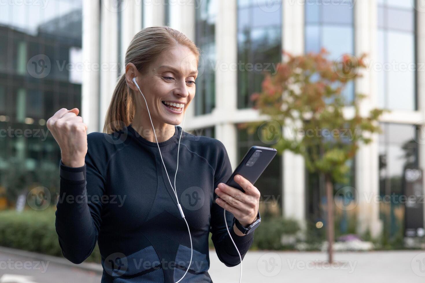 A cheerful woman celebrates a moment of victory or good news outdoors, listening to music with earphones from her smartphone. The backdrop showcases urban greenery and modern architecture, evoking a sense of a work-life balance. photo