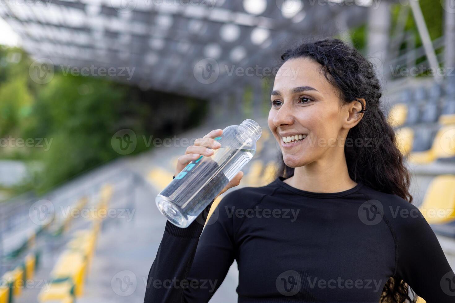 Smiling athletic woman staying hydrated with a water bottle at a sports stadium. Health and fitness lifestyle concept captured outdoors. photo