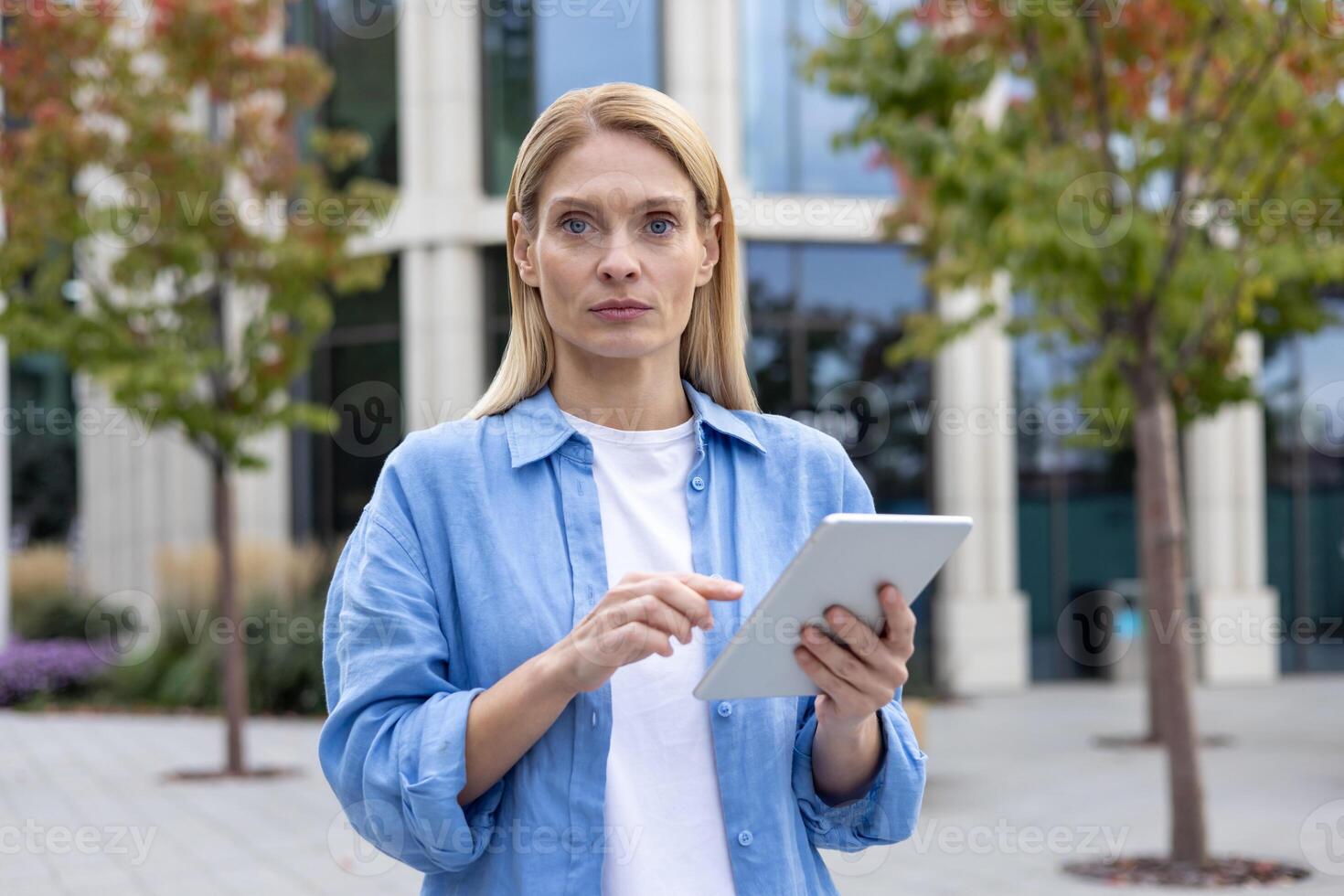 A focused professional woman stands outside on a cloudy day, using a digital tablet in front of a contemporary office building, embodying modern business and technology. photo