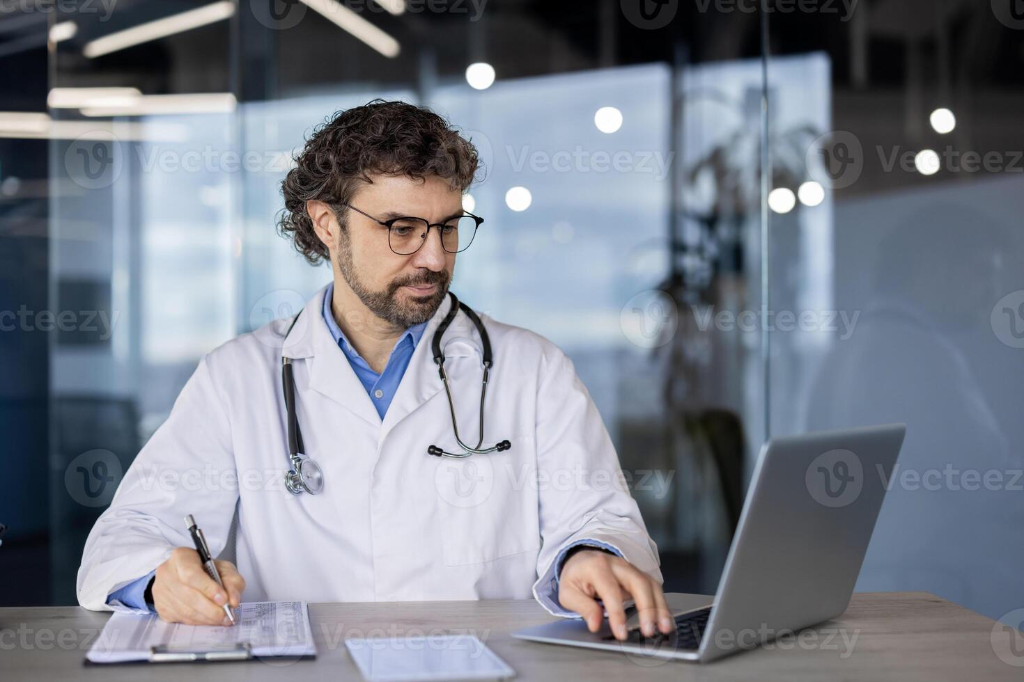 A professional male doctor in a lab coat using a laptop and writing notes on a clipboard in a modern healthcare setting. photo