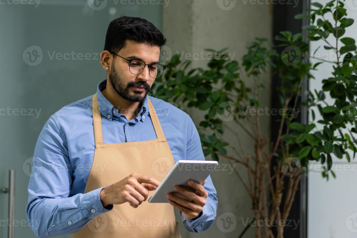 un dedicado hombre con un barba y lentes se enfoca en su tableta mientras vistiendo un casual delantal, encarnando emprendimiento y planta cuidado. foto